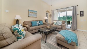 A living room with beige sofas, a coffee table, and a view of greenery through large windows. - Rent the Perfect 4 Bedroom Villa on Windsor Palms Resort, Orlando Villa 4988 (Kissimmee)