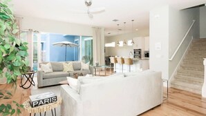 Ein modernes Wohnzimmer mit einem grauen Sofa, einem weißen Sofa und einem kleinen Tisch mit einem 'PARIS'-Schild. Rechts befindet sich eine Treppe und im Hintergrund ein Küchenbereich.
