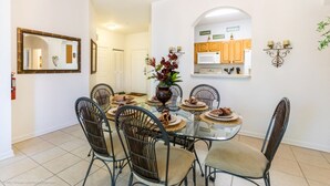 A dining area with a glass table surrounded by wicker chairs, a vase with flowers, and a kitchen in the background. - Rent a Luxury Apartment on Windsor Hills Resort, Minutes From Disney, Orlando Apartment 3207 (Kissimmee)