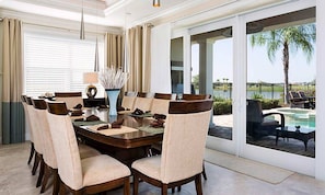 A dining area with a round table, cushioned chairs, a vase with white flowers, and a view of a pool and palm trees through large sliding glass doors. - Rent Your Own Orlando Villa With Large Private Pool on Reunion Resort and Spa, Orlando Villa 4561 (Kissimmee)