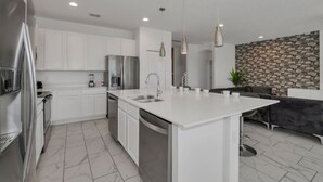 Une cuisine moderne avec des meubles blancs, un grand îlot central doté d’un évier, des appareils en acier inoxydable, un éclairage suspendu et un mur en pierre en arrière-plan.