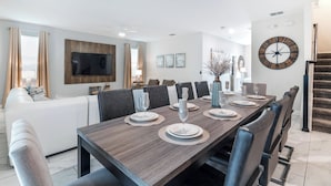 Une salle à manger moderne avec une longue table en bois, dressée pour six personnes, entourée de chaises aux couleurs sombres. Un téléviseur à écran plat est fixé au mur, au-dessus de la table.