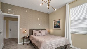 A bedroom with a bed, two bedside lamps, a ceiling light fixture, a door, a window with blinds, and a painting on the wall.