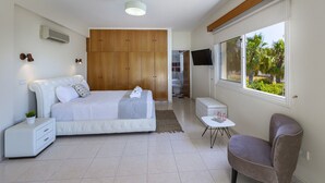 Un dormitorio moderno con una cama grande, un armario de madera, una mesita pequeña, una silla y un televisor.