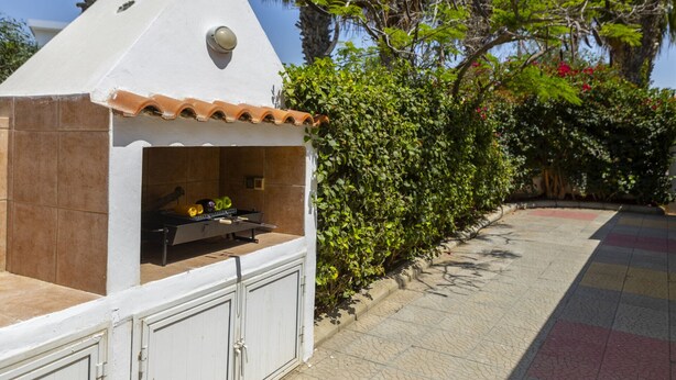 Zona de parrilla exterior pequeña con revestimiento blanco y techo marrón, rodeada de follaje y un camino de baldosas.