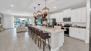 Une cuisine spacieuse dotée d’un grand îlot central, de meubles blancs, d’appareils électroménagers en acier inoxydable et d’un luminaire suspendu moderne.