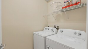 A laundry room with two front-loading washers and dryers, a white wall, and a shelf above the dryers holding a basket and a box. - Your Family Will Love the 1st Class Amenities at Your Private Villa on Windsor Island Resort, Orlando Villa 5044 (Kissimmee)