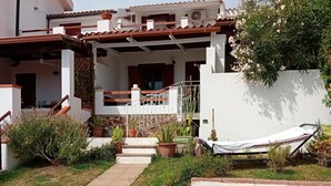 Una propiedad residencial con un camino pavimentado que conduce a un porche cubierto, rodeada de plantas en macetas y una hamaca.
