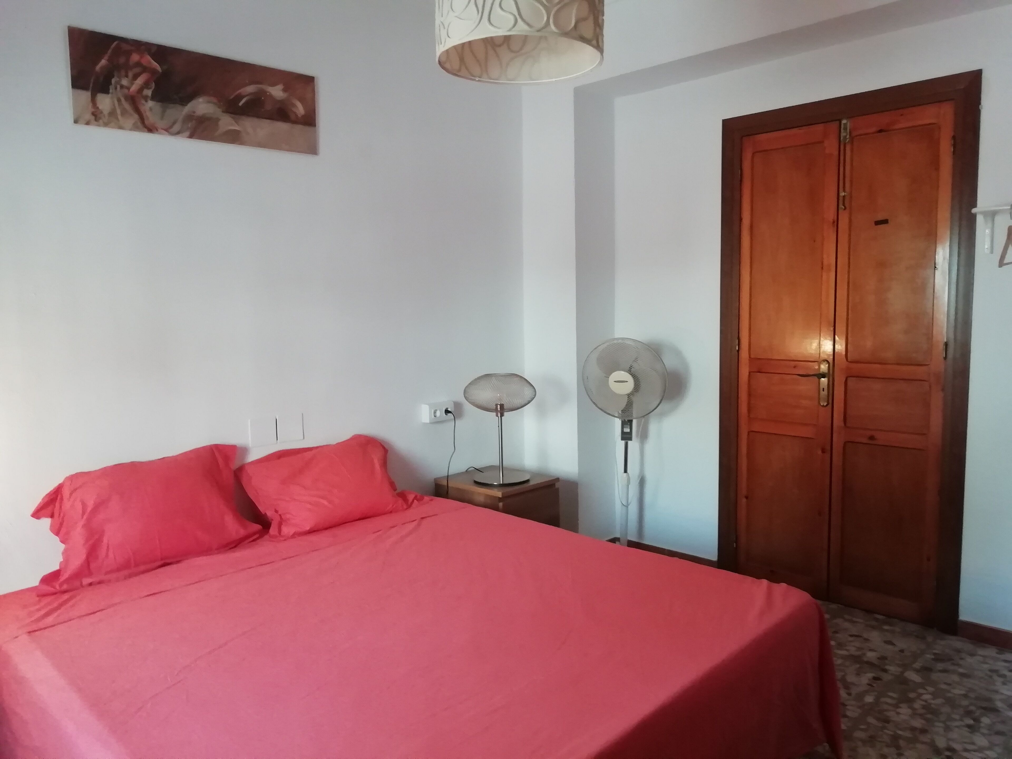 A room with a bed, a wooden door, a wall-mounted fan, and a ceiling light fixture.