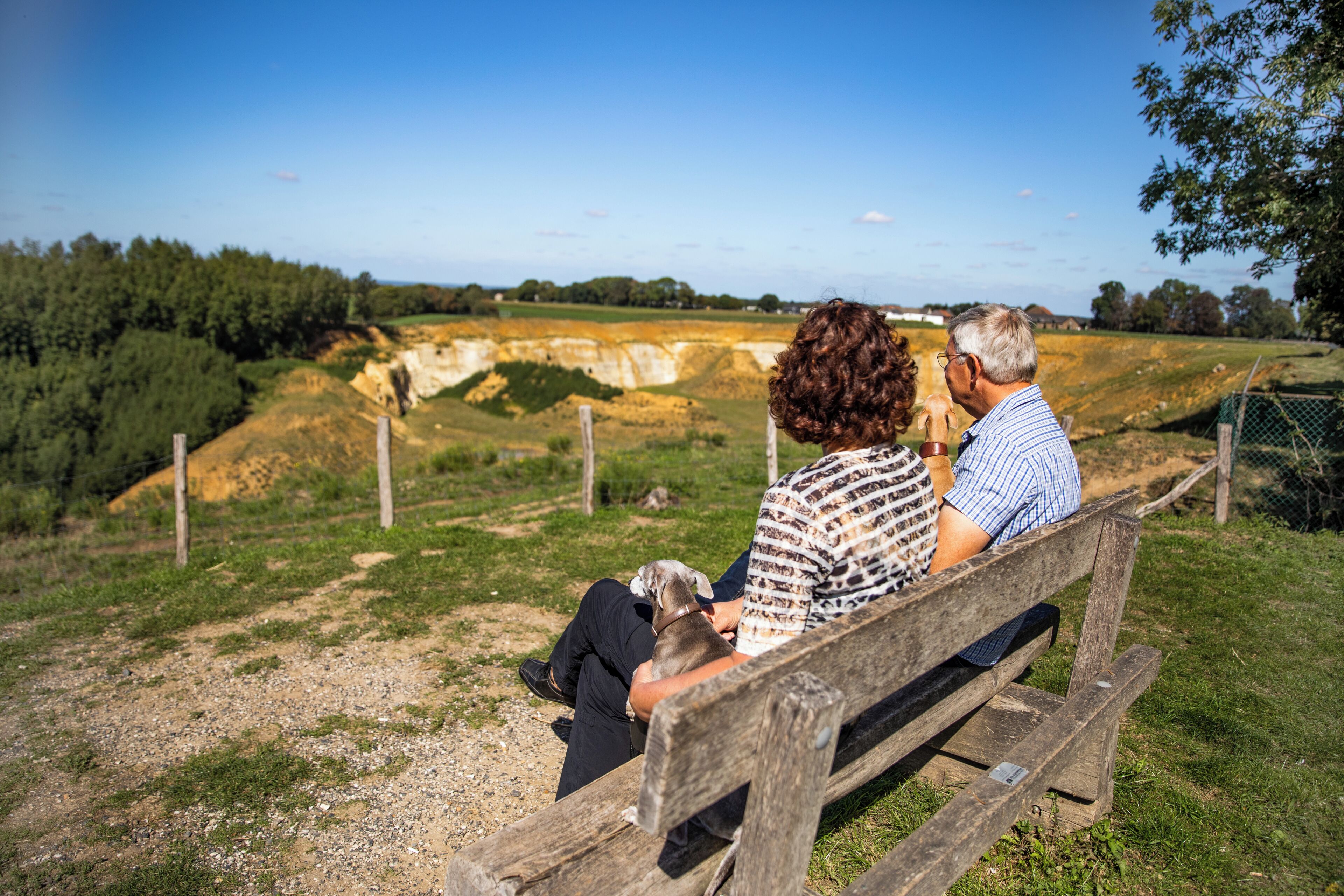 A couple sitting on a wooden bench overlooking a natural landscape with a fence and trees.