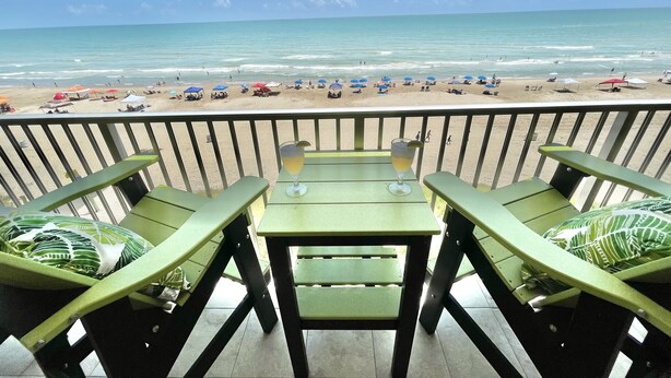A balcony with a view of the beach, featuring a table with two chairs and a glass of drink.