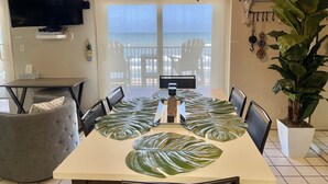 A dining area with a table set for four, a ceiling fan, a television, and a view of the ocean through a sliding door.