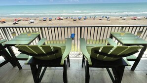A balcony with green chairs and tables overlooking a beach with umbrellas and people.