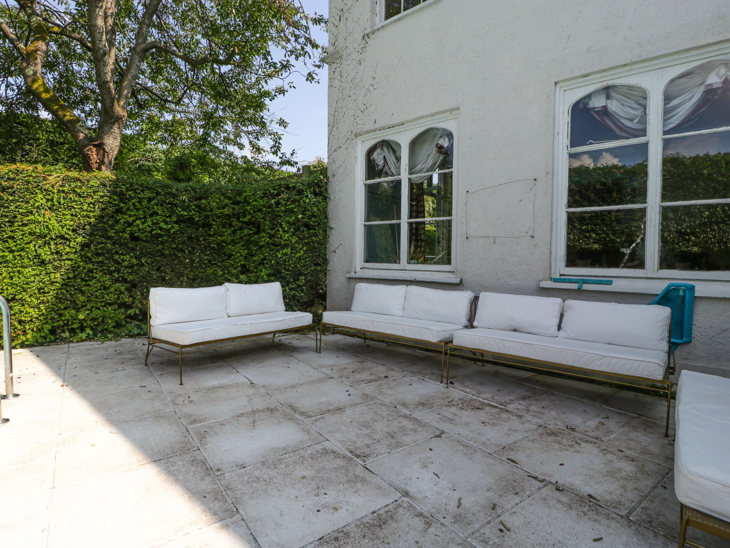 A patio with three white sofas and a table, a hedge, and a building with arched windows.