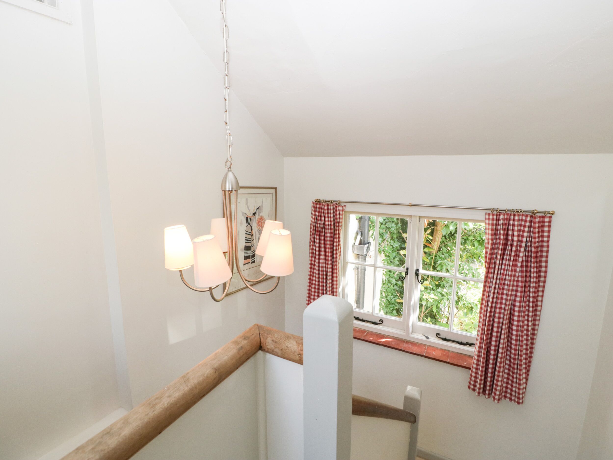 Een trap met een houten leuning, een kroonluchter met vier lampen en een raam met rood-witte geruite gordijnen.