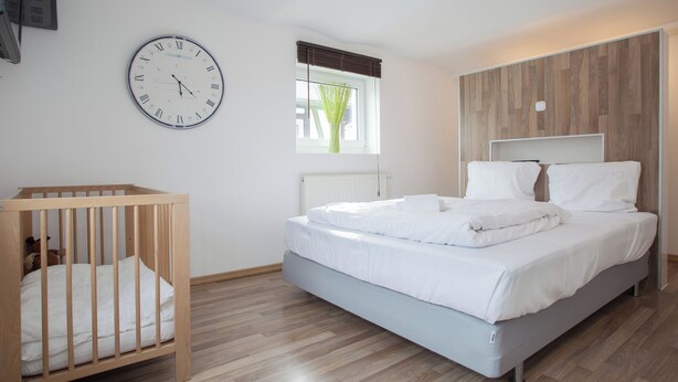 Een slaapkamer met een houten ledikant, een bed met witte beddengoed, een wandklok en een raam met zonwering.