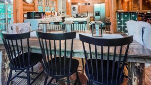 Un intérieur rustique en bois, avec un coin repas comprenant une table et quatre chaises, une cuisine équipée d’appareils modernes et un lustre orné de plusieurs ampoules.