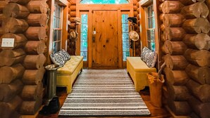 L’intérieur d’une cabane en rondins, avec une porte centrale, deux bancs garnis de coussins, un étendoir suspendu et un tapis à motifs.