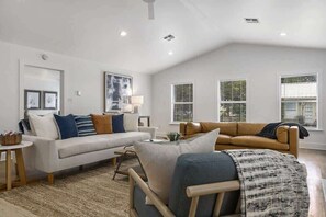 Ein geräumiges Wohnzimmer mit einem weißen Sofa, einem braunen Sofa und einem Holzstuhl. Zwei Fenster mit Jalousien, ein Bild an der Wand und ein kleiner Tisch mit einer Schale sind ebenfalls zu sehen.
