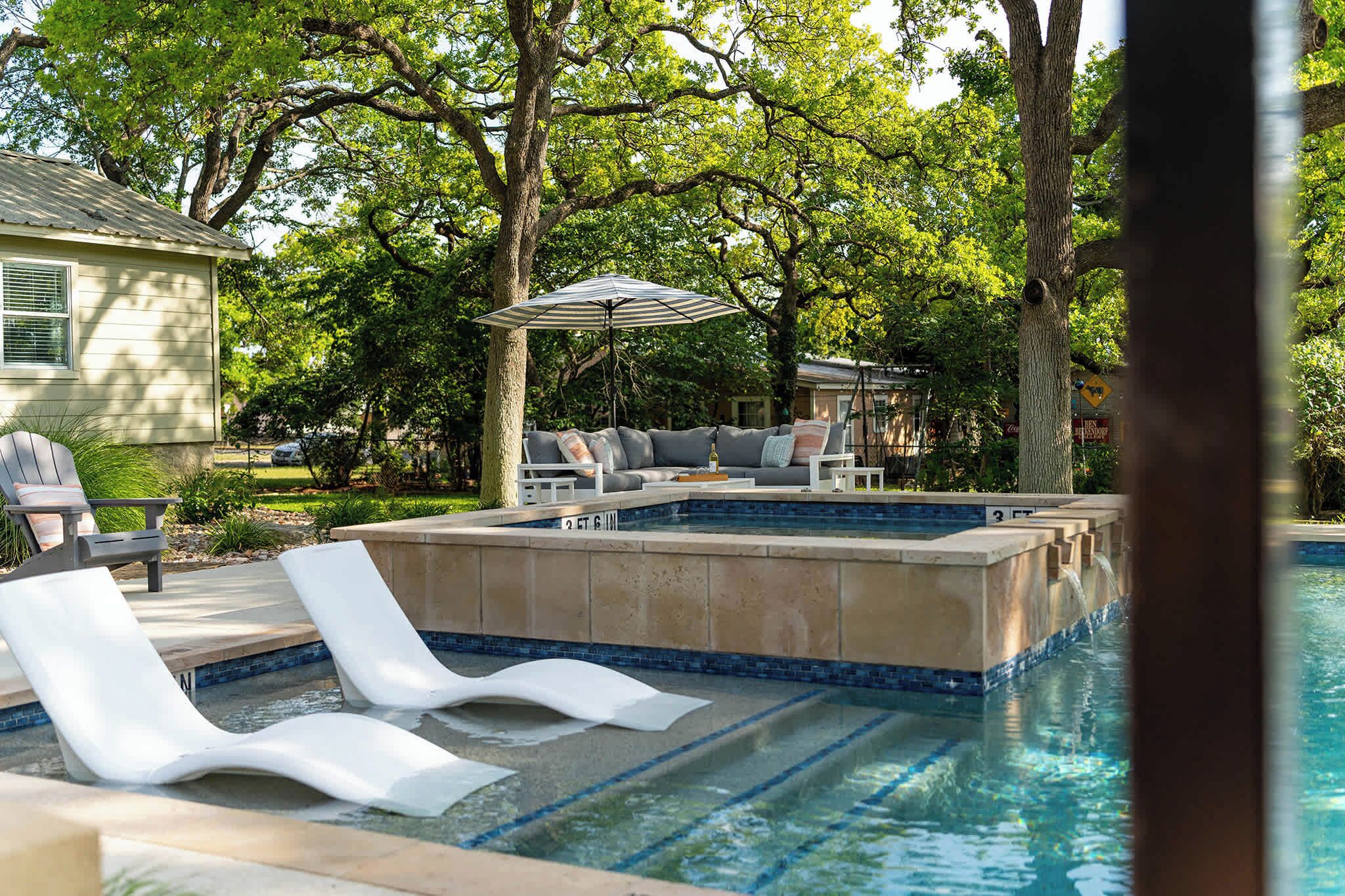 Un área de piscina con sillas de descanso, un espacio de estar con una mesa y sillas, y una sombrilla.