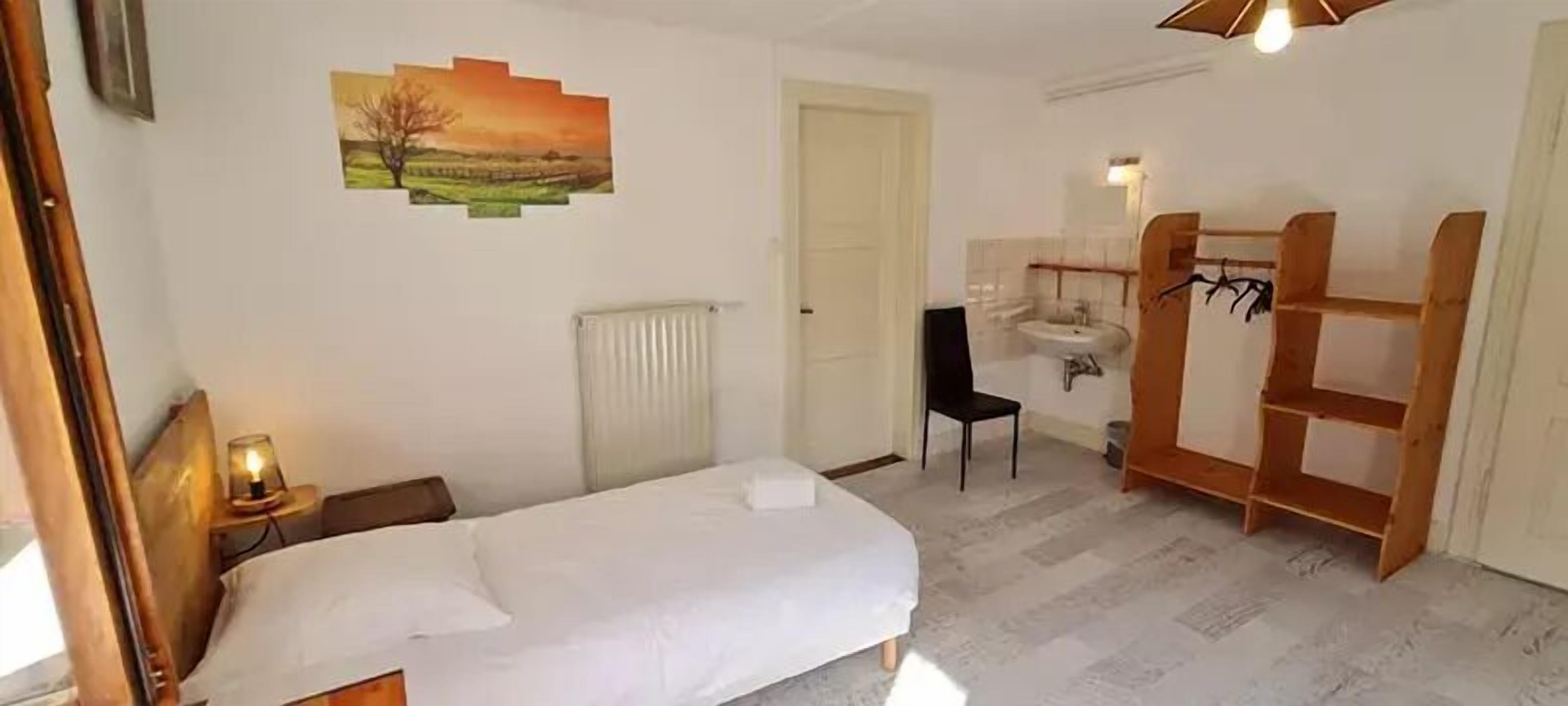 A bedroom with a bed, a radiator, a chair, a wooden shelf unit, and a painting on the wall.