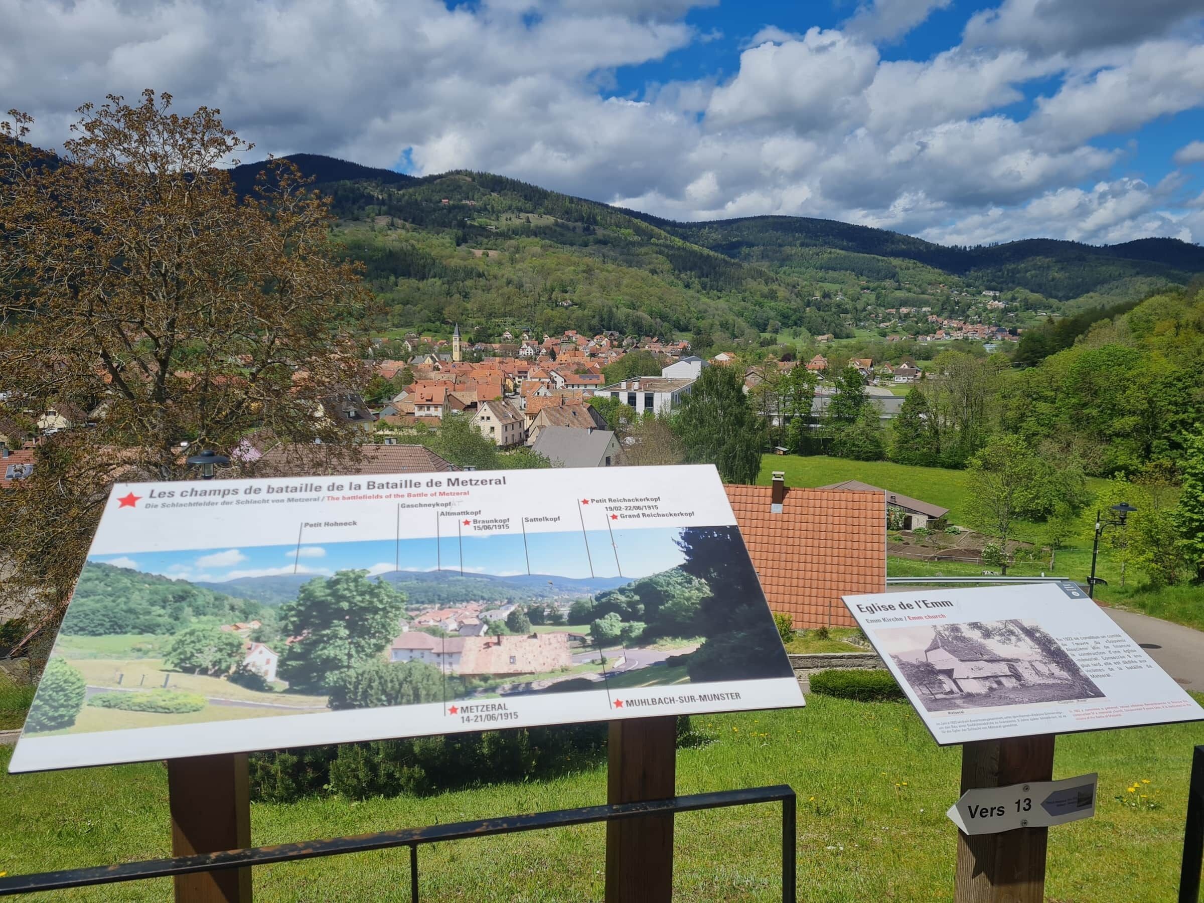 A historical battlefield site with informational signs, a village in the background, and a mountainous landscape.