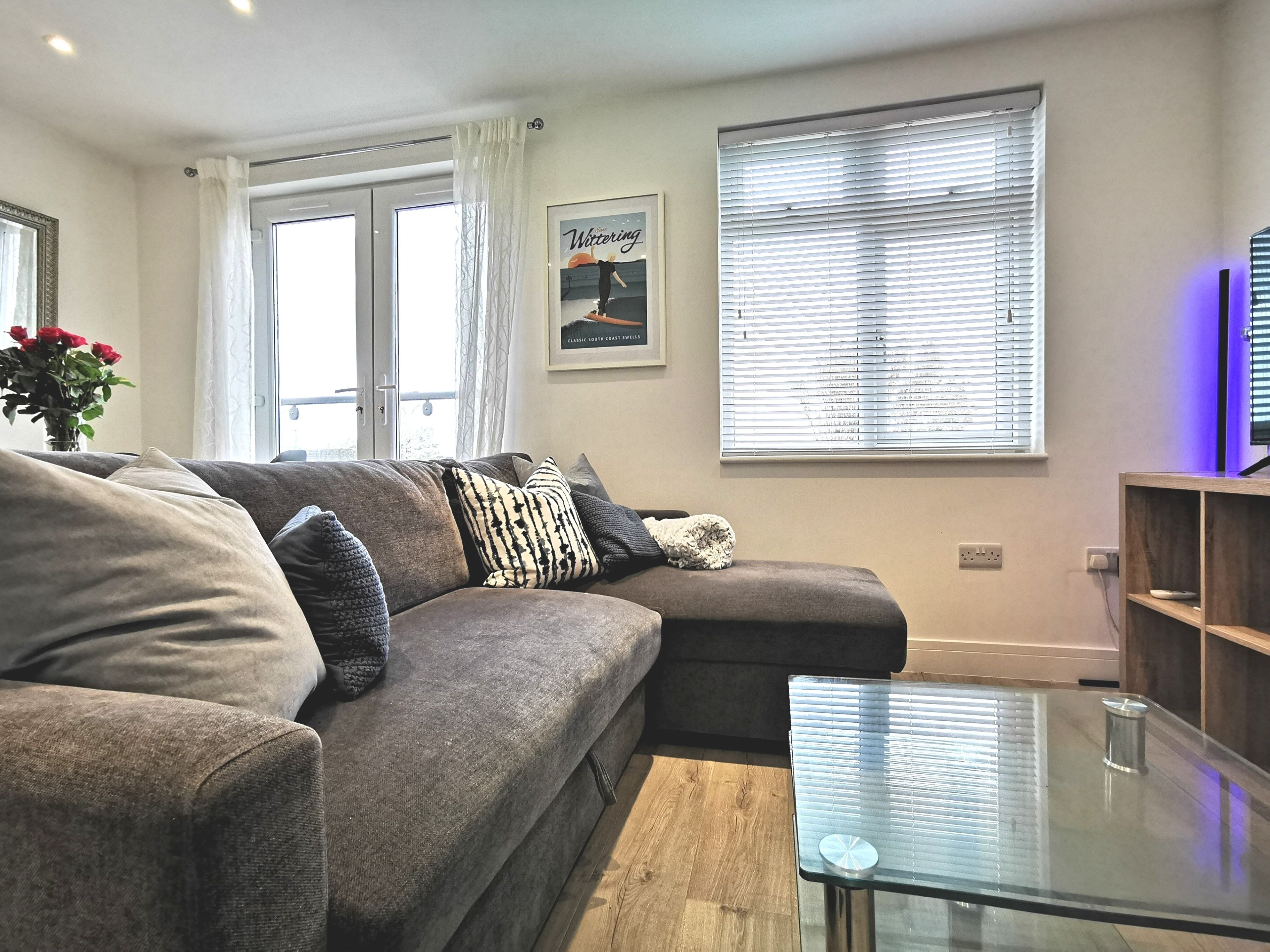 A modern living room with a grey sectional sofa, a glass coffee table, and a large window with blinds.