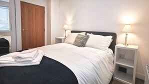 A neatly made bed with white linens and a black accent, two bedside tables with lamps, a wooden door, and a window with blinds.