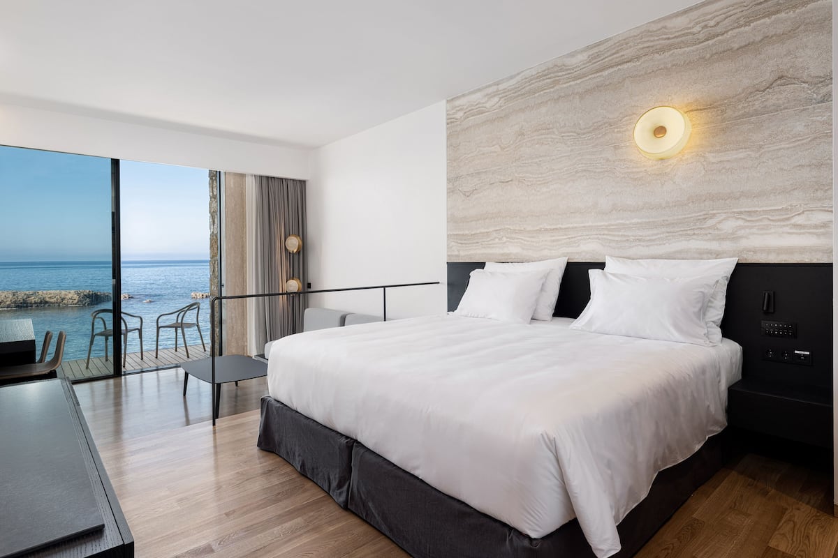 A modern bedroom with a large bed, a desk, and a view of the sea.
