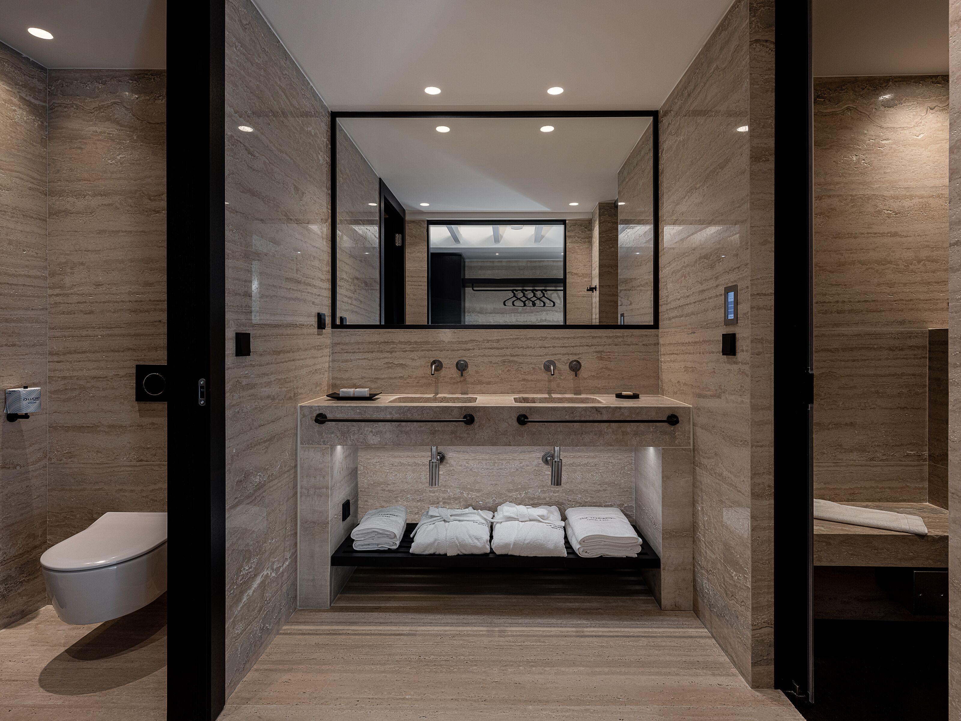 A modern bathroom with a marble wall, a large mirror, a sink with two faucets, and a towel rack.