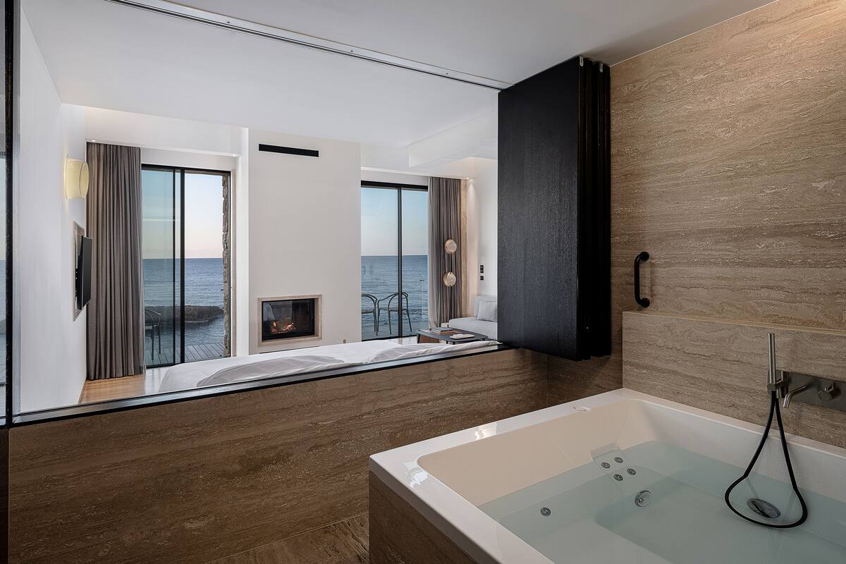 A modern bathroom with a bathtub, a glass shower enclosure, and a view of the sea through large windows.