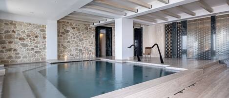 A modern indoor swimming pool with stone walls, a wooden ceiling, and a chair near the entrance.