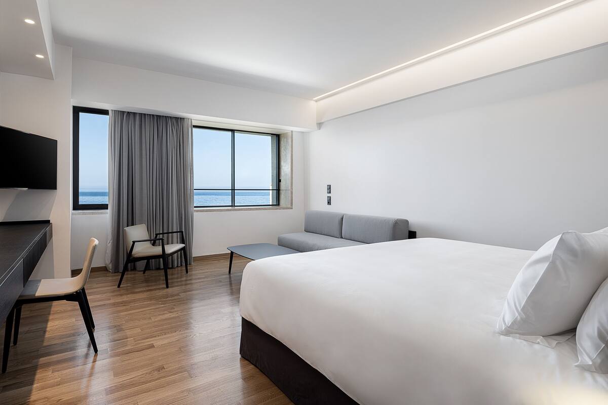 A modern hotel room with a large bed, a sofa, a desk with a chair, a TV mounted on the wall, and a window with a view of the sea.