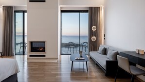 A modern hotel room with a large sliding glass door to the sea, a fireplace, a sofa, and a dining area.