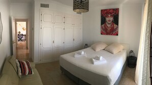 Ein Schlafzimmer mit einem großen Bett, einem Sofa mit gemustertem Überwurf, einem Bild an der Wand und einer Tür, die in einen Flur führt.