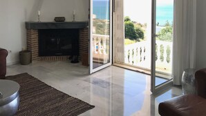Ein großzügiger Wohnraum mit Kamin, einer Kristallleuchtre und Blick auf das Meer durch eine Schiebeglastür.