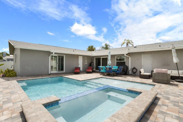 Un jardin avec une piscine rectangulaire, un patio couvert aménagé avec du mobilier d’extérieur, et une maison à l’extérieur grise et à la porte blanche.
