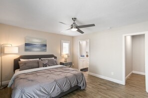 Une chambre à coucher comprenant un lit, un ventilateur de plafond, une lampe de chevet, une table de nuit, un tableau, une fenêtre avec des stores, une salle de bain avec un miroir et une porte donnant sur une autre pièce.