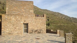 A stone building with a door and a signboard, surrounded by a stone-paved area and a hillside in the background.