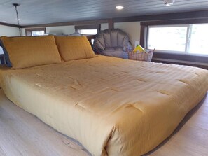 A large bed with a beige quilt, a gray chair, and a basket of items on a wooden floor.