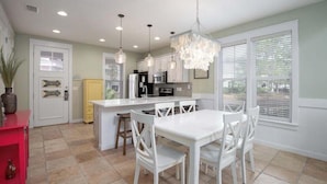 A kitchen with white cabinetry, a central island with seating, pendant lighting, and a view through large windows. - Mermaid Manor - with 6-seater Golf Cart! (Miramar Beach)