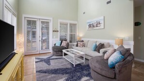 A living room with a sofa, coffee table, and a TV. There are two doors with glass panels and a ceiling fan. - Mermaid Manor - with 6-seater Golf Cart! (Miramar Beach)