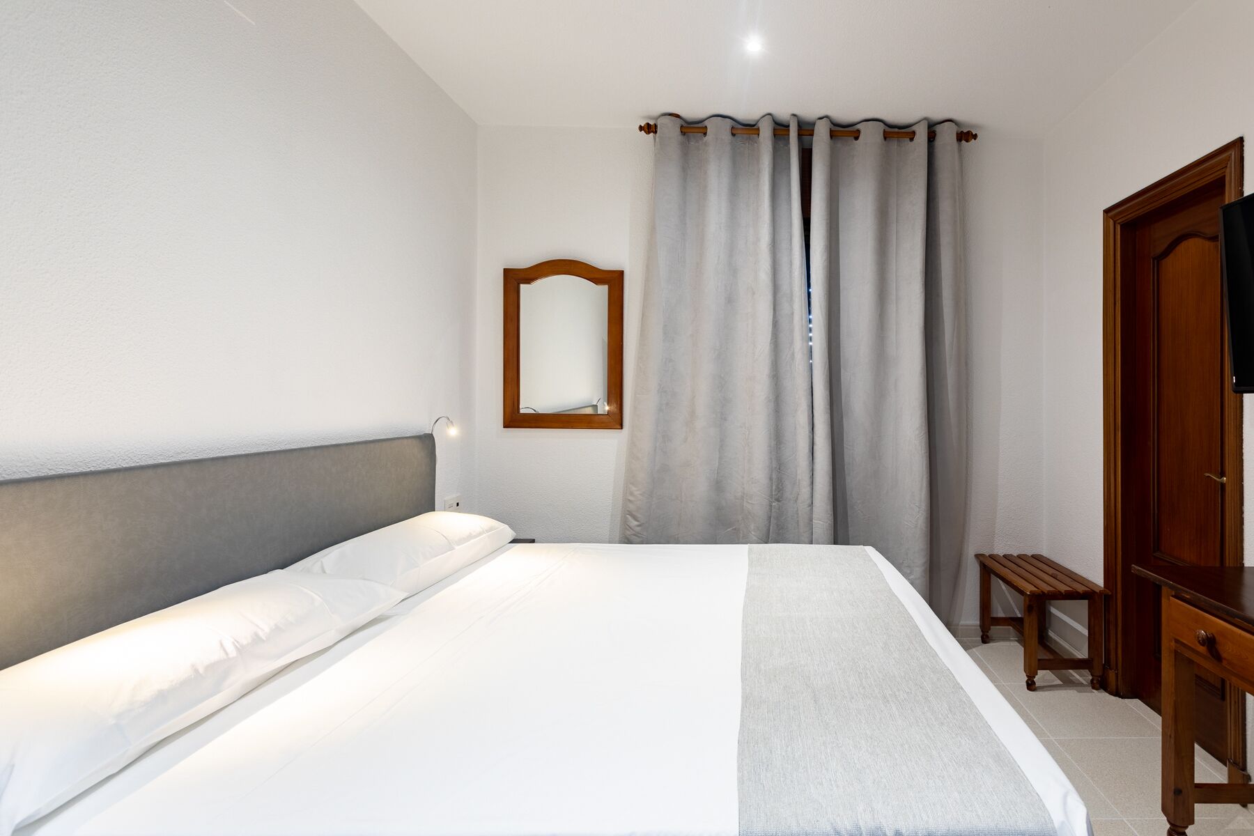 A neatly made bed with a grey headboard, a wooden bench, and a wooden wardrobe in a simple, clean room with white walls and a grey curtain.