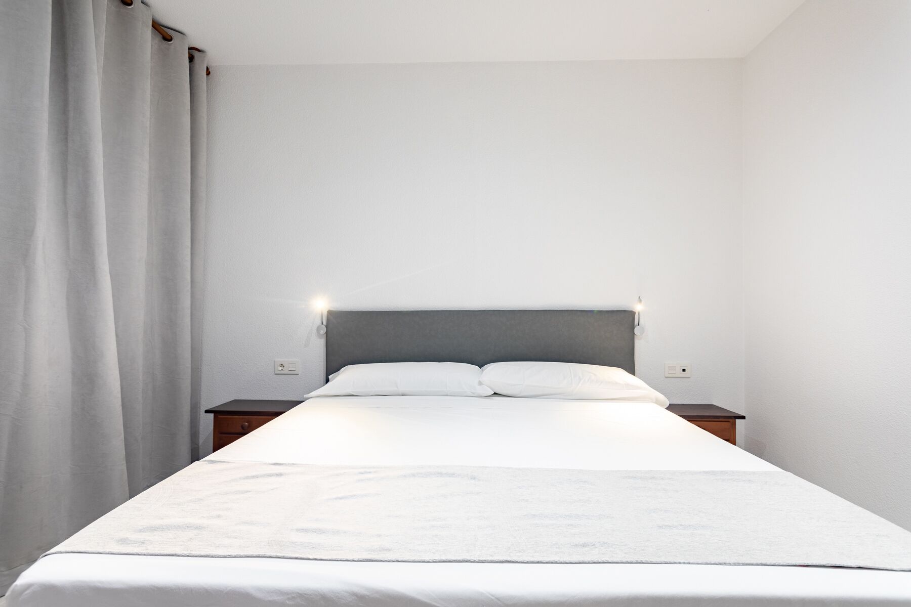 A minimalist hotel room with a single bed, white bedding, and a simple headboard.