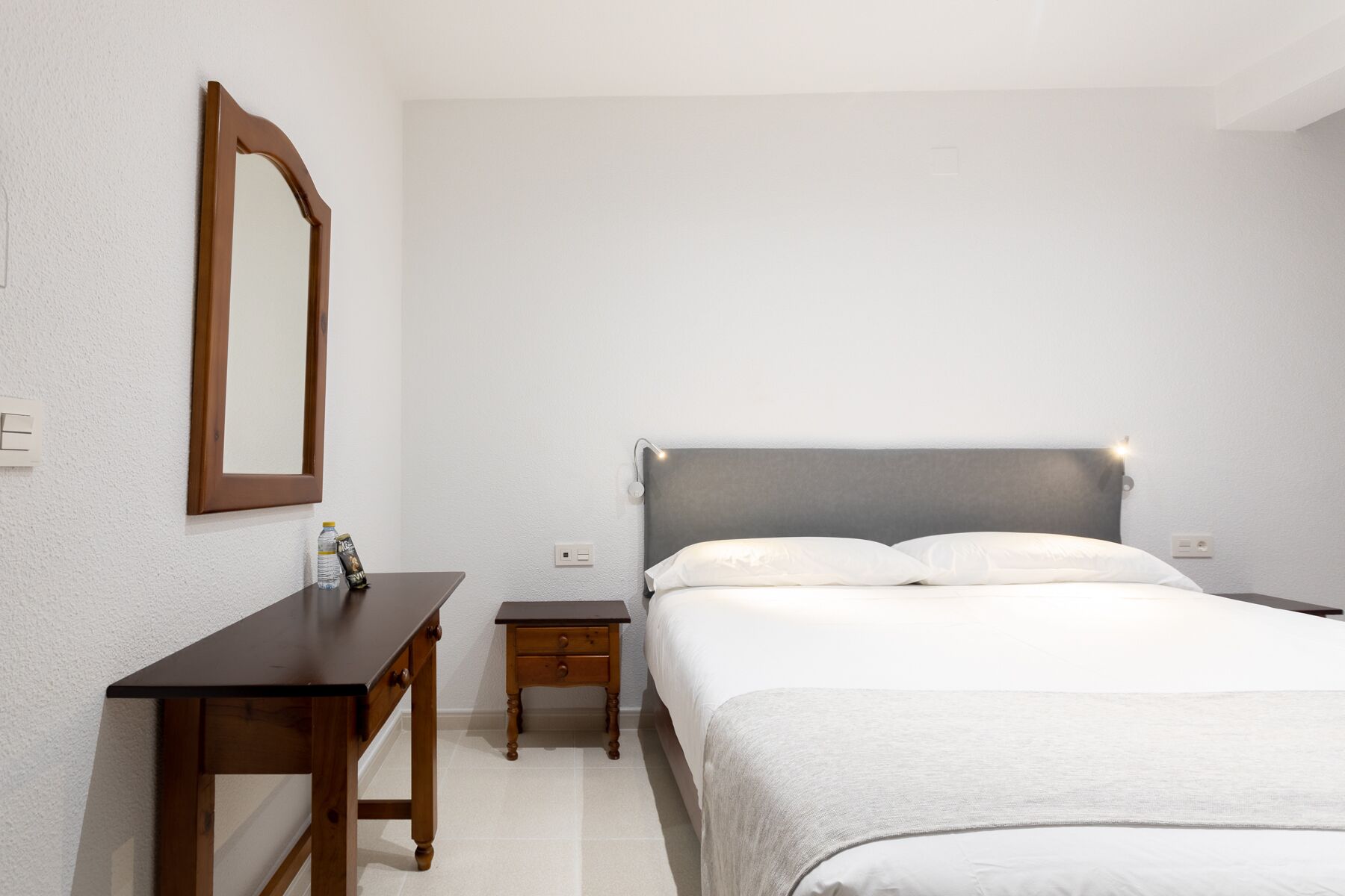 A bedroom with a bed, a bedside table, a nightstand, a mirror, and a wall socket.