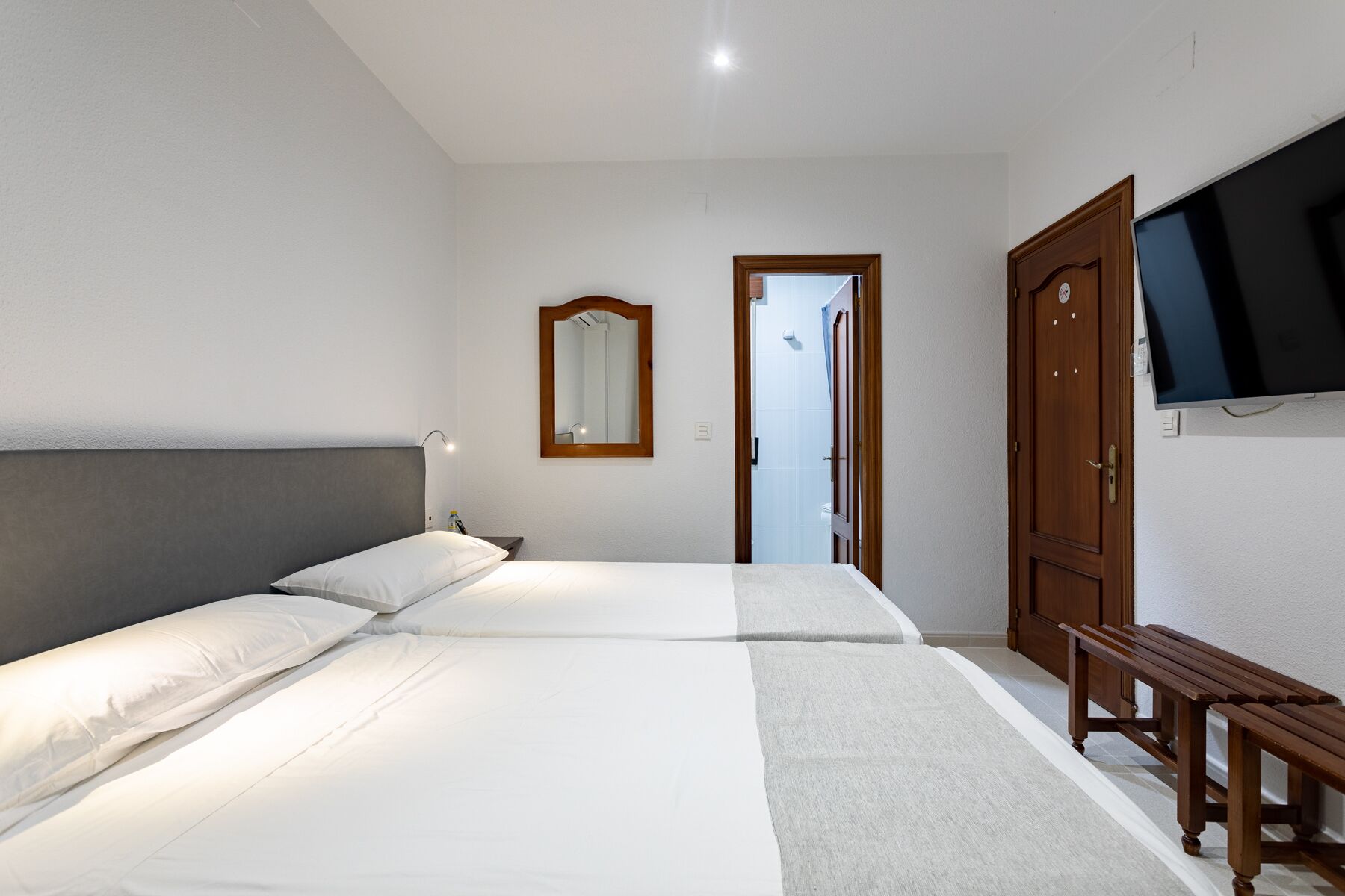 A hotel room with two beds, a wooden bench, a TV mounted on the wall, and a mirror above the bed.