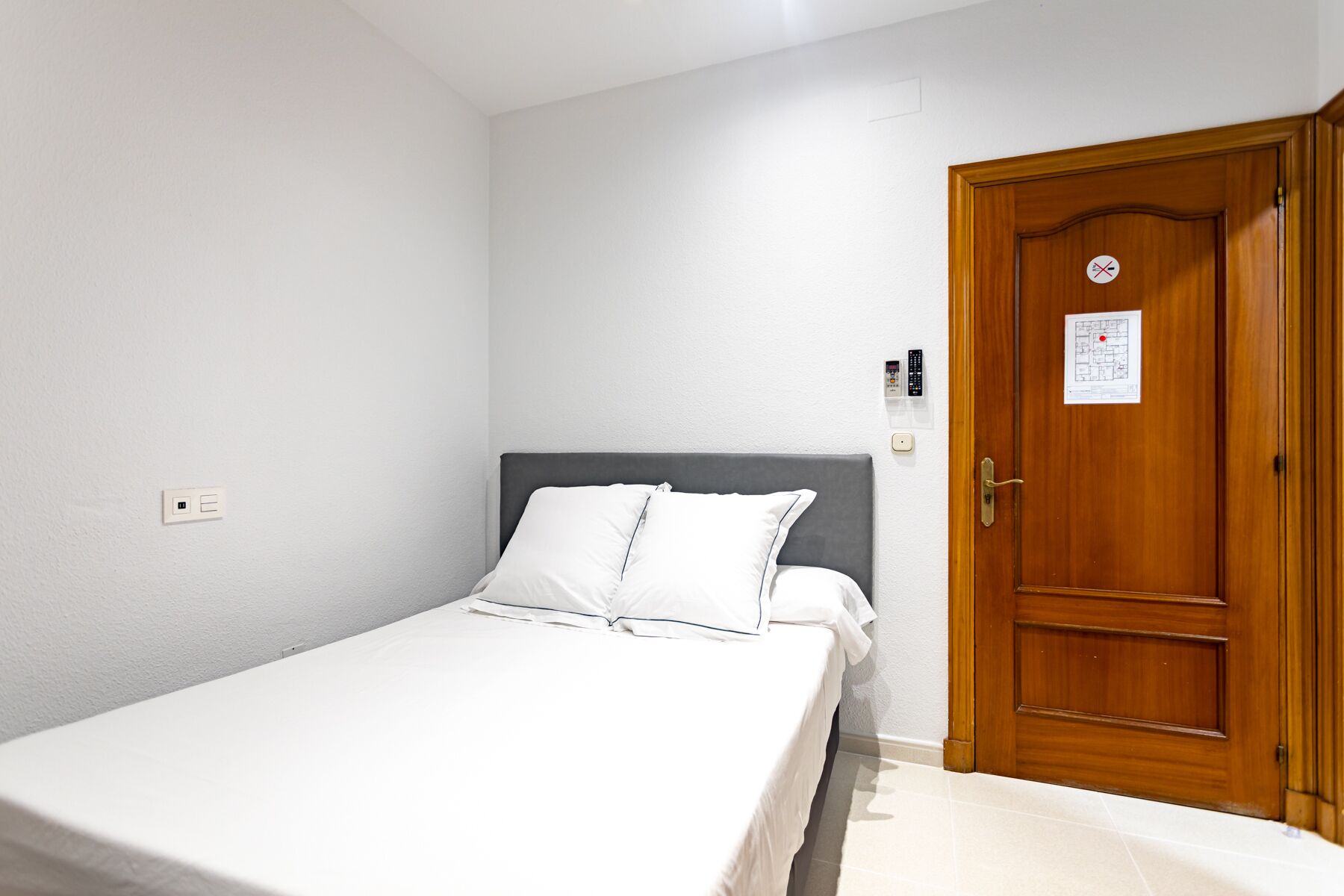 A single bed with white bedding in a room with a wooden door and a light switch on the wall.