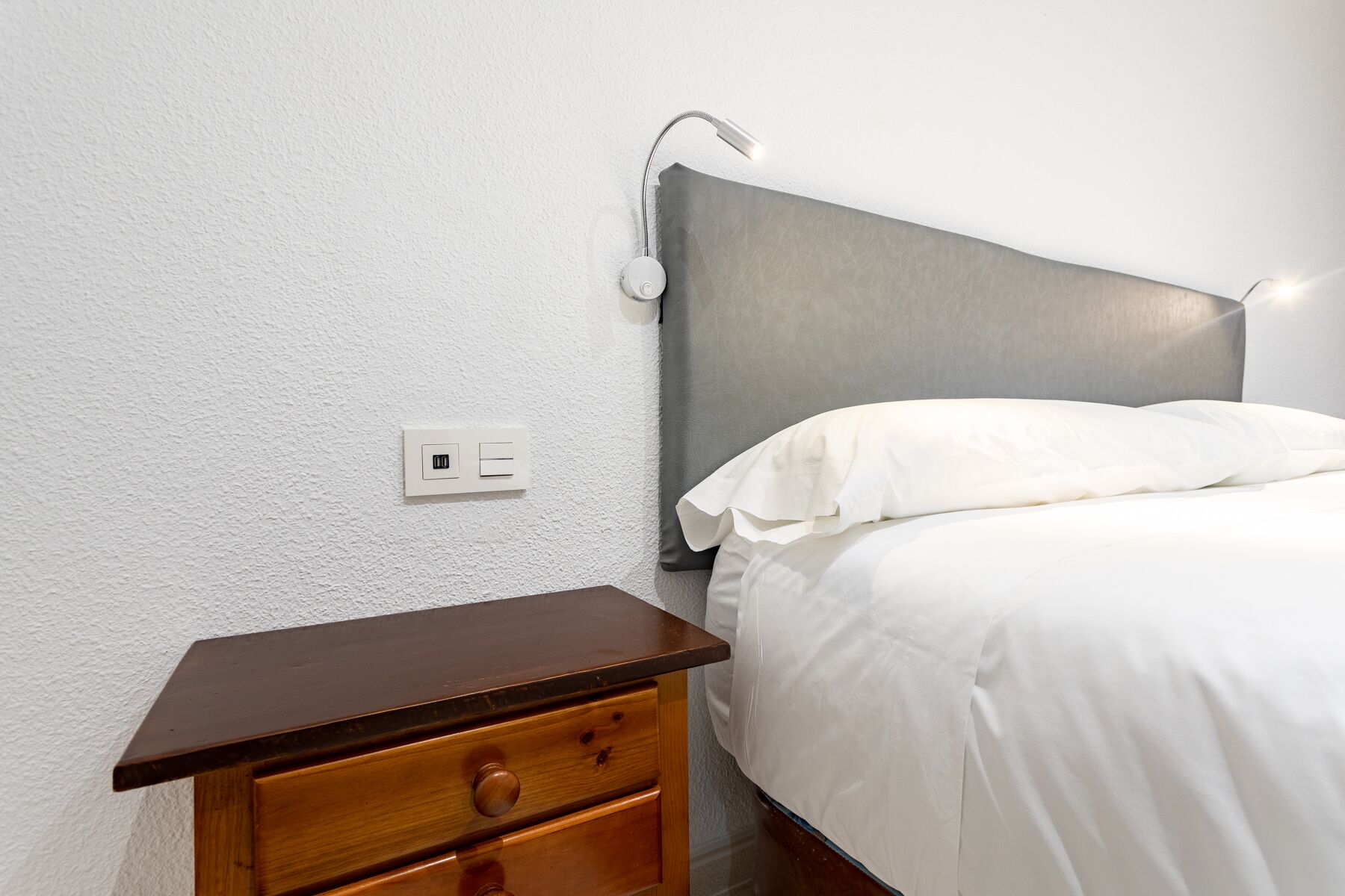 A bedroom with a wooden nightstand, a bed with white bedding, a gray headboard, and a wall-mounted lamp.