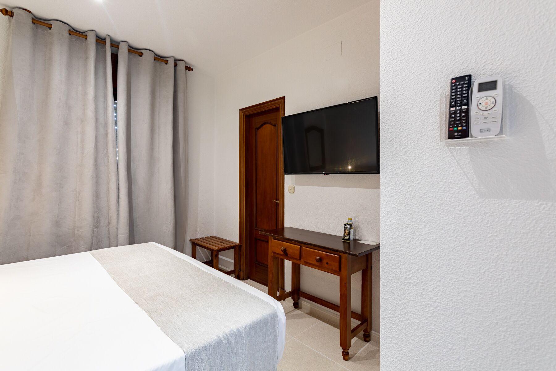 Een hotelkamer met een bed, een bureau, een aan de muur bevestigde televisie en een bedieningspaneel aan de wand.