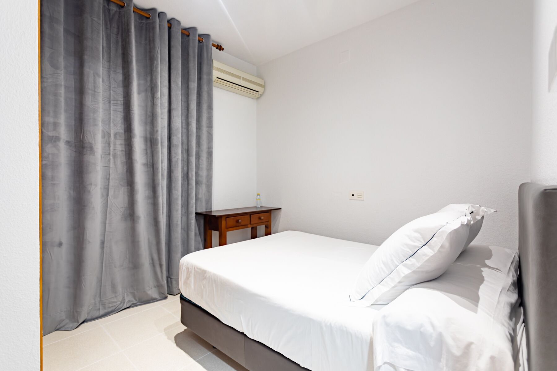 Une chambre à coucher avec un lit, une table de chevet, une bouteille d’eau et un climatiseur fixé au mur.